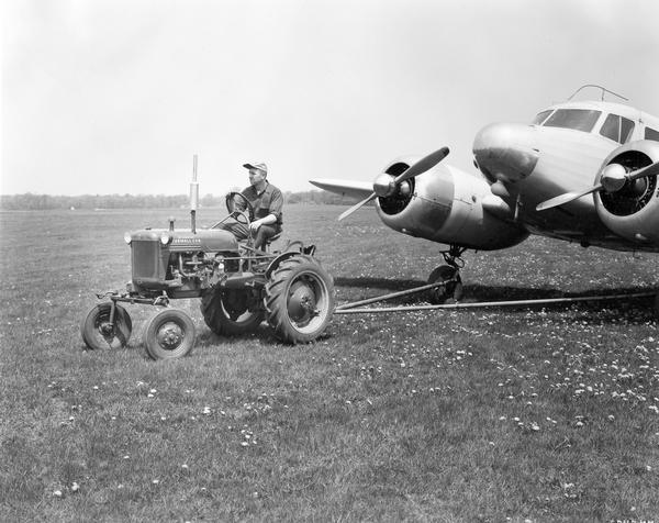Airport worker using a McCormick Farmall Cub tractor equipped with towing bar and special towing bracket to tow a small twin-engine airplane at the Elmhurst Airport.