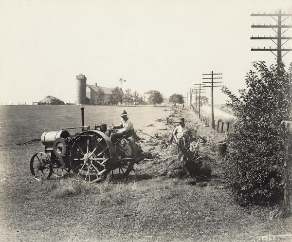 Two farmers are using an International Harvester Titan 10-20 HP tractor to remove tree stumps along the fence line of a farm near a road. Electrical poles are running along the fence line. In the background are farm buildings, a silo and a windmill.