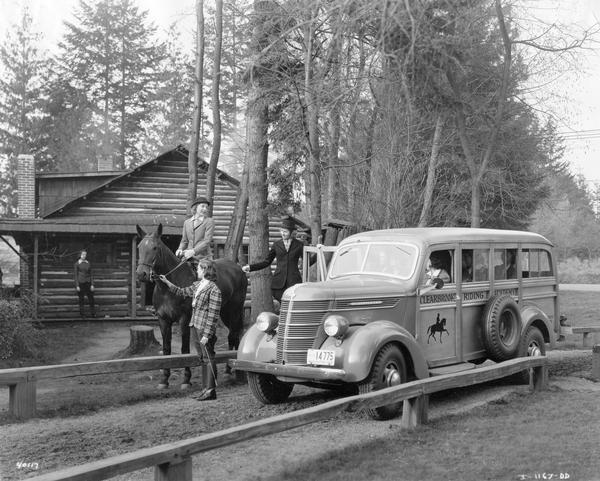 Female students and a horse next to, and inside of, an International D-15-M station wagon ("woody") used for transportation at the Clearbrook Riding Academy. A log cabin is in the background.