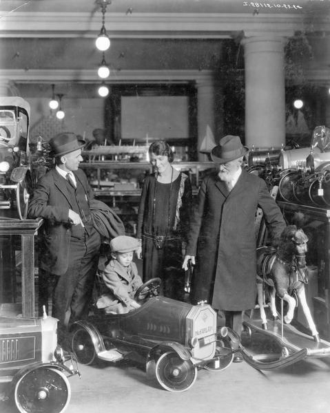 Boy sitting in a "McFarlan" toy car as two men and a woman are looking on. The group appears to be in a toy store or toy department, with other toy cars, a rocking horse and toy sailboats.