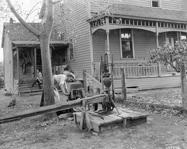 Farm woman operating a McCormick-Deering portable gasoline engine. The engine is driving a pump to draw water from a well into a homemade trough. Another woman is watching from the porch of a house in the background.
