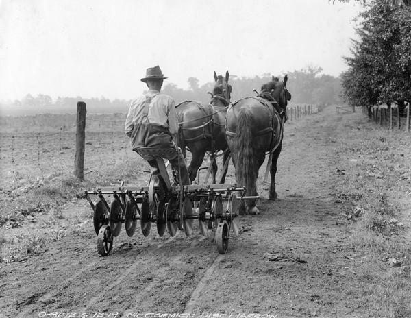 Farmer riding down a dirt path on a horse-drawn McCormick disc harrow on transport wheels.
