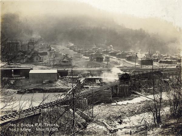 Small steam engine hauling empty cars over a bridge along a narrow gauge railroad track. Houses in a residential neighborhood are in the background. The train is on the no. 2 bridge for the no. 2 mine. Benham was a "company town" created by International Harvester for the workers of the Wisconsin Steel Company. Wisconsin Steel was a subsidiary of International Harvester and operated coal mines at Benham.