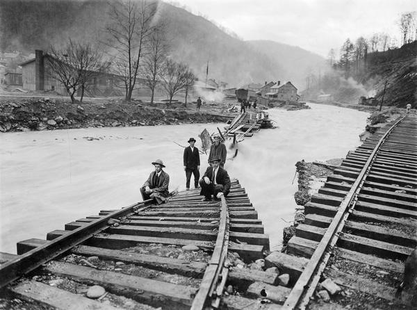 Men standing on the exposed portion of a flooded railroad track as a swiftly moving torrent of water is rushing past. Benham was a "company town" created by International Harvester for the workers of the Wisconsin Steel Company. Wisconsin Steel was a subsidiary of International Harvester and operated coal mines at Benham.
