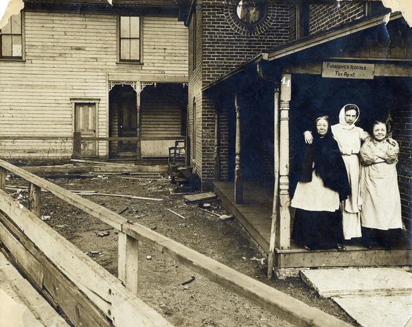 View over board fence towards three women standing on the porch of a boarding house, possibly in Dayton, Ohio. The porch is adjacent to a yard littered with debris. A handwritten sign above their heads reads: "Furnished Rooms For Rent, 44." The photograph was taken by or for International Harvester's Agricultural Extension Department, and was likely intended to illustrate urban living conditions.