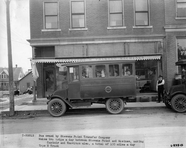 International Model H bus loaded with passengers and parked in front of a store selling furniture and "talking machines" (phonographs). The bus was owned by the Stevens Point Transfer Company. According to the original caption, the bus "makes two trips a day between Stevens Point and Mosinee, making Casimir and Knowlton also, a total of 100 miles a day."