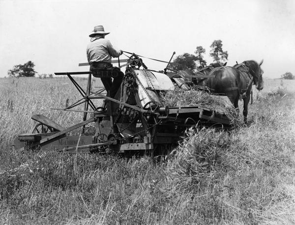 Farmer harvesting grain with horse-drawn McCormick harvester and wire grain binder built in 1876. The scene is likely a historical re-enactment - possibly created in conjunction with International Harvester's "reaper centennial" celebration. Original caption reads: "The wire binding attachment consisted mainly of two steel fingers which moved back and forth and twisted a wire band around each sheaf of grain. This machine did the cutting and binding in a single operation, requiring but one man to drive the team. McCormick built and sold 50,000 wire binders between 1877 and 1885."