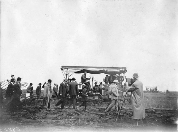Men in suits and hats follow along as a man plows with a Holt crawler tractor at the Winnipeg tractor contest in Canada. Another man in the foreground is preparing to photograph the proceedings. The tractor was built by Holt Manufacturing Company of Stockton, California.