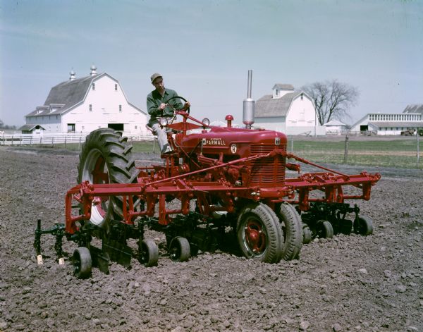 View towards a farmer in a field with a stage I McCormick Farmall Super H tractor and attached four-row cultivator. Farm buildings are in the background.