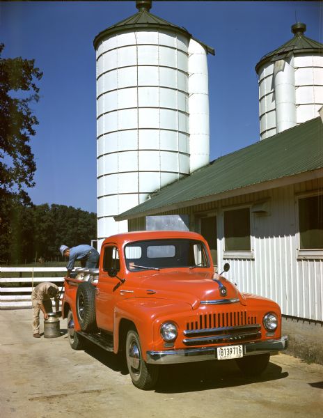 Color photograph of two men loading milk cans into the back end of International L-120 pickup truck. In the background are two grain silos.