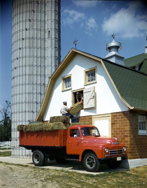 Color photograph of two men unloading bales of hay from an International L-150(?) truck into a barn.