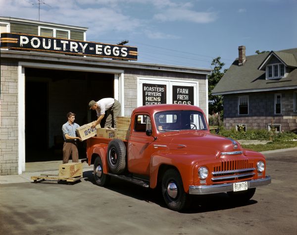 Two men are loading boxes of eggs into the back end of an International L-120 pickup truck in front of a poultry store. Produced from 1949-1953, approximately 32,125 L-120 models were made.