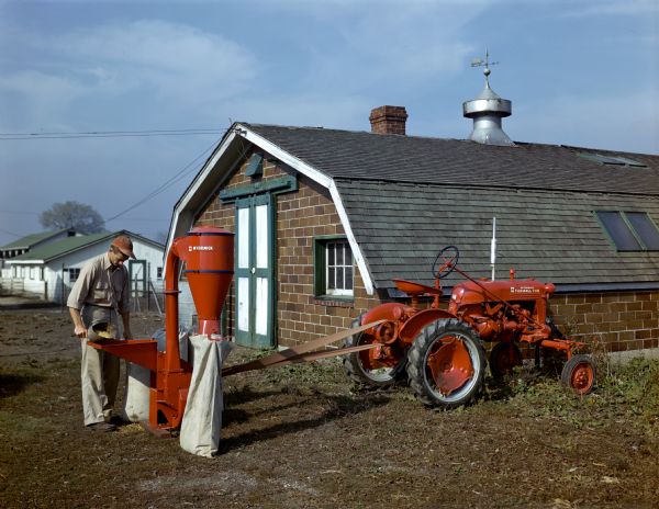 A farmer is standing and pouring grain into a McCormick belt-driven hammer mill powered by a McCormick Farmall Cub tractor.