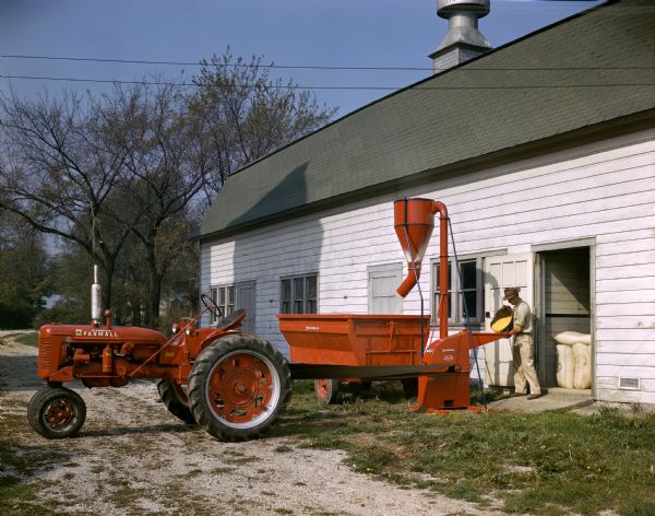 View towards a farmer standing at an open barn door pouring corn into a belt-driven McCormick hammer mill powered by a Farmall C tractor.
