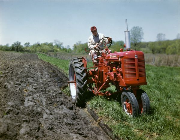 View of a farmer plowing a field along a fence line with a McCormick Farmall Super C tractor.