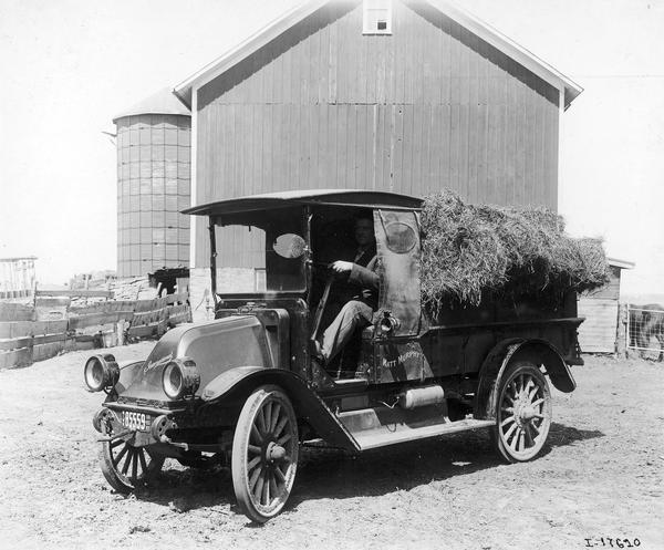 Farmer in an International Model H truck loaded with hay. A barn and silo are in the background. The H was produced from 1915-1923.
