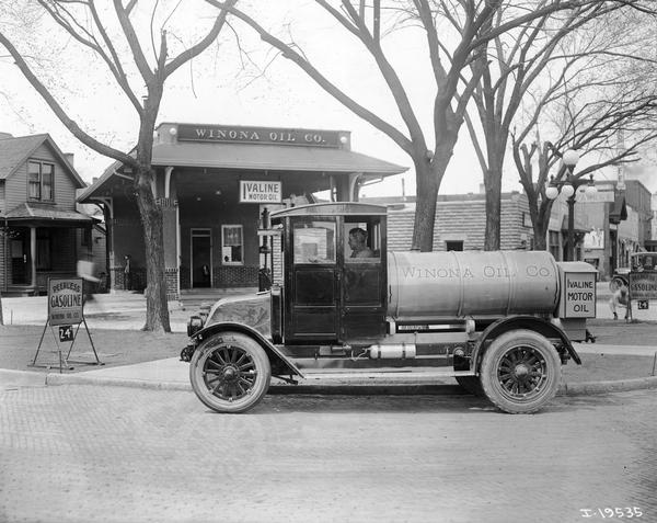 Driver's side view of a man sitting in the driver's seat of an International Model H oil truck owned by the Winona Oil Company. The truck is parked outside the Winona Oil Company gas station. There are signs advertising "Peerless Gasoline" and "Ivaline Motor Oil."