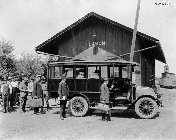 Passengers lined up to board an International Model H bus in front of a railroad depot at Lamoni(?).