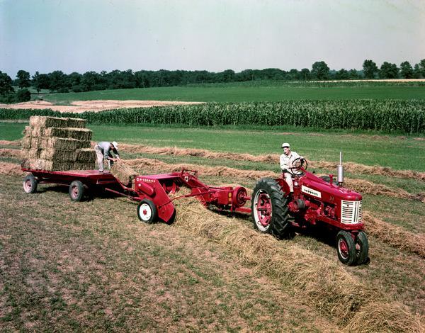 Color photograph, slightly elevated, of two farmers baling hay with a McCormick Farmall 350 tractor, a McCormick baler and a wagon.