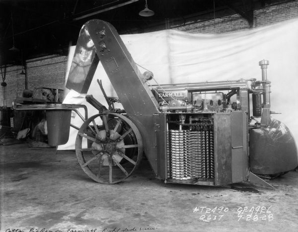 Engineering photograph of an experimental cotton picker attached to a Farmall tractor. A white backdrop is along the brick wall in the background.