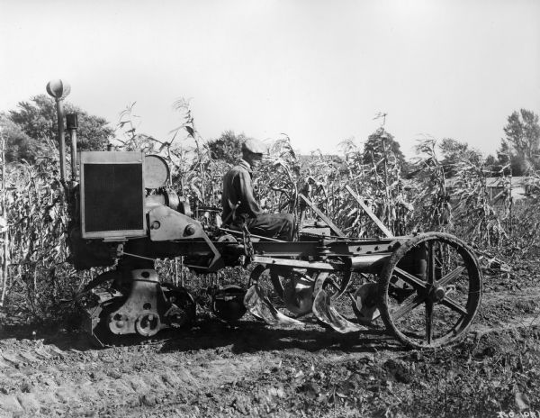 Engineering photograph of a farmer field testing an experimental motor cultivator with crawler treads. Original caption reads: "The motor cultivator with plow raised for transportation... The plow was raised by a crank having a sleeve which slipped over the winch connection."