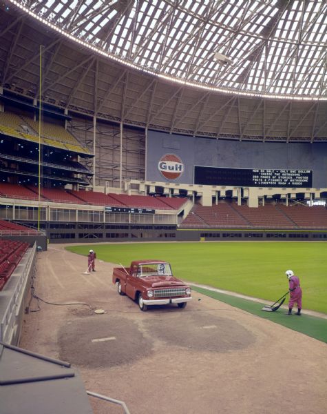 Color photograph of Houston Astrodome groundskeepers dressed in "moon suits" getting the field ready for the 1966 baseball season. An International D-Line pickup truck used by the groundskeepers is parked in foul territory by the bullpen.