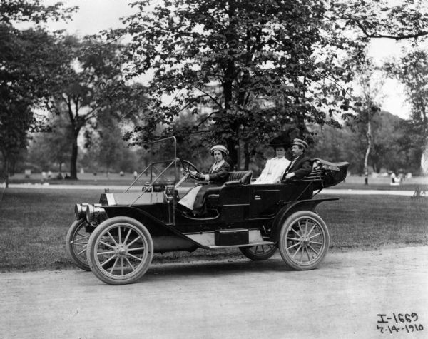 Woman and two passengers driving an International J-30 passenger car with its top down through a city park. Also known as the IHC 30, the J-30 was produced from 1910 to 1911. A total of 1,105 were built. Standard colors were a dark blue body and primrose yellow running gear.
