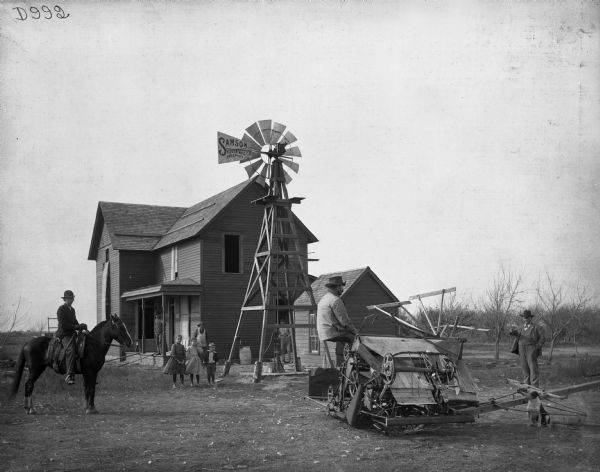Farm family standing in front of their house, while on the right a man is sitting on a Deering binder, and two men on the left and right are looking on. At the center of the scene is a windmill with the sign "Sampson Tower Mfg. Co. Freeport, ILL." The house may still be under construction.