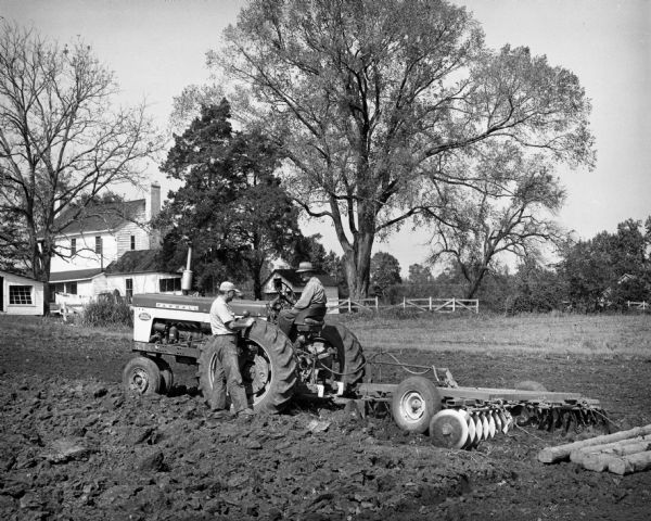 Farmer on a Farmall 560 tractor with disk harrow pausing to talk to another man in a field in front of a farmhouse.