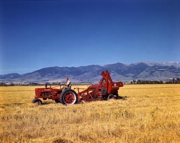 Farmer harvesting hay with a McCormick-Deering Farmall H tractor and no. 64 combine (harvester-thresher) set against a background of mountains.