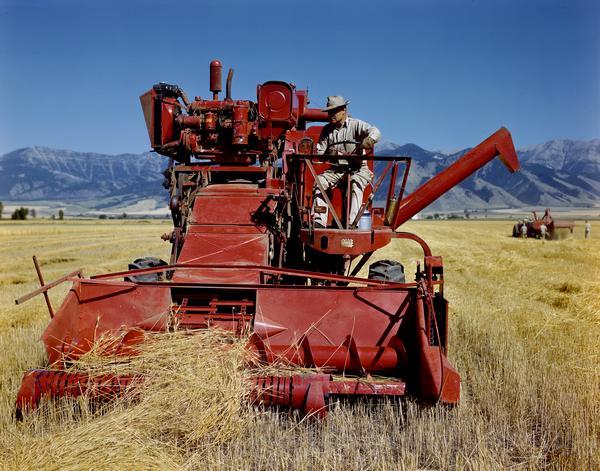 Farmer harvesting wheat with a McCormick combine (harvester-thresher) against a mountainous background. The combine is using a windrow pickup head and may be experimental.
