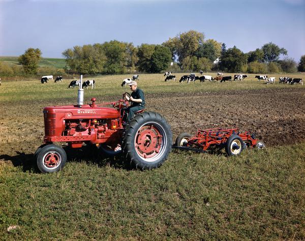 Left side view of a farmer on a McCormick Farmall M tractor with plow in a field. Cows are grazing in the background.