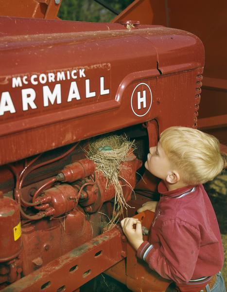 Young boy inspecting a robin's nest on the engine of a Farmall H tractor. Original caption reads: "Spring-time on the farm. Even though his father uses this tractor every morning for filling silage wagons, mama robin persisted in building her nest and laying her eggs next to the engine block. The eggs hatched and the young robins eventually grew and flew."