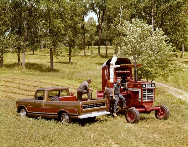 Color advertising photograph showing two men in the field refueling an International 966 Hydro tractor with a 1972 International 1010 pickup. The tractor has a forage chopper and chopper box attached.