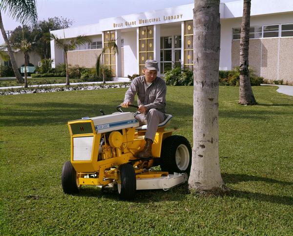A male worker is mowing the grounds of the Belle Glade Municipal Library with an International Cub Cadet 125 tractor and mower. The Cub Cadet 125 was produced at the Louisville works beginning in 1968.