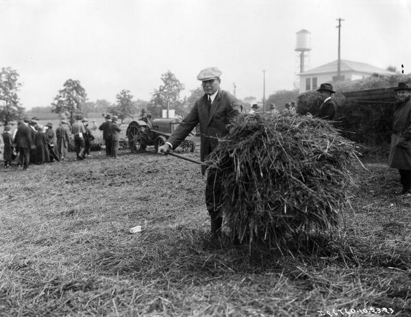 Alexander Legge (1866-1933) with a pitchfork full of hay at a Thresher demonstration at International Harvester's Hinsdale farm. Men are milling around a tractor in the background. Legge was president of International Harvester Company from 1922 to 1929. He was vice chairman of the War Industries Board during World War I and later appointed chairman of the Federal Farm Board by President Hoover in 1929. He became president of International Harvester again in 1931 and remained in that position until his death in 1933.