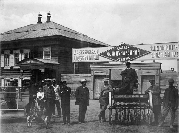 Several men and a young child pose with a cultivator and other implements in front of a Russian agricultural equipment dealer. The dealer may have been an International Harvester or McCormick dealer.