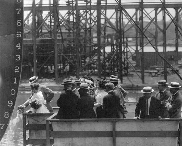 Christening ceremony for the S.S. <i>Harvester</i>. The ship was built for the Wisconsin Steel Company, a subsidiary of International Harvester. The 545-foot all steel freighter was built by the American Shipbuilding Company using the "Isherwood" system of design. Attending the ceremony were: Miss Marie Smith, daughter of the Captain (breaking the bottle); F.R. Gadd of the Chicago WSW; H.F. Hughes, International Harvester Traffic Department; Captain William Smith of Algonac; Chief Engineer Wallace Tomey of Cleveland; R.H. Hibble of San Francisco; Lloyds' representative John French of Cleveland; Marine Architect Robert Logan; Lindsey Wallace of the American Shipbuilding Company; Mrs. Wallace Tomey and daughter.