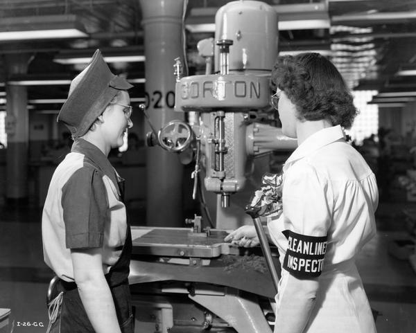 Two women inspect a drill press at an International Harvester factory. Original caption reads: "Dust prevention and cleanliness is so important as a factor in accuracy on some of the torpedo production jobs that a nurse in one of the dispensaries also acts as cleanliness inspector. She makes regular daily inspections of the department checking cleanliness,  and has authority to stop any employee in his/her work and instruct them to brush up any dust or dirt. Here she is shown pointing out metal shavings to an employee and asking that they be removed."