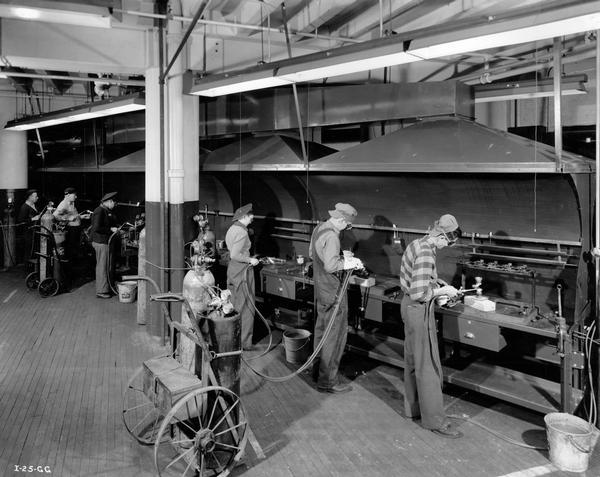 Male and female workers producing torpedo parts at an International Harvester factory. Original caption reads: "A battery of acetylene torches is here shown used in the coppersmith department to braze, solder and sweat torpedo parts. These operators have completed special courses in this work which requires careful and precise operation."