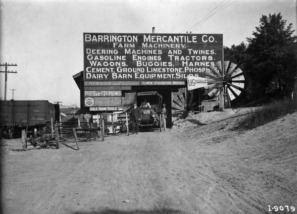 Storefront of Barrington Mercantile Company, a rural agricultural implement dealer. A variety of signs indicate equipment sold, including Deering machines and twines; Rock Island plows; Emerson farm machinery; New Stoughton wagons; and Gale farm tools. An International Highwheeler is parked in front of the building.
