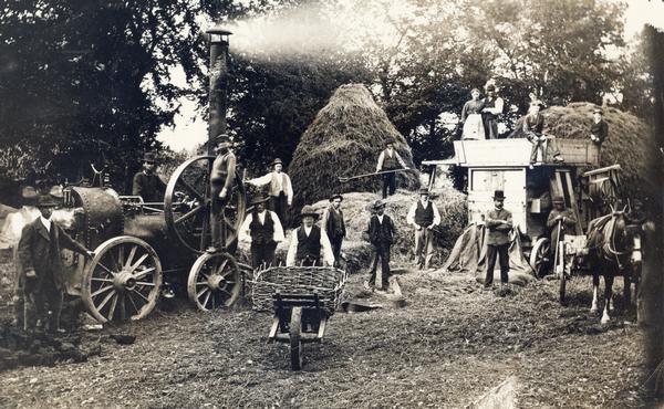 Men and women pose in the middle of threshing operations with steam engine, horse-drawn wagon and a wheelbarrow equipped with a homemade basket. A handwritten note on the back of the print reads: "threshing in the 1870s; Miss O'Hanlin, the Holt, Hook, Hants(?), England."