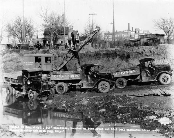 Three International HS-54(?) dump trucks haul dirt from an excavation site. The trucks were owned by R.J. Blackburn, Inc. and Arrow Hauling Co., Inc.