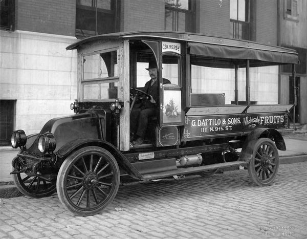 Man sitting in an International Model G delivery truck along street curb. The truck was owned by G. Dattilo & Sons, wholesalers of fruits, and featured a custom body built by Sam V. Aspromonte.