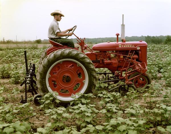 Right side view of a young man riding a Farmall Super C tractor with attached cultivator in a field.