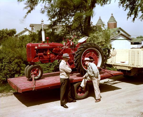 Slightly elevated view of two men talking next to a trailer loaded with a Farmall Super C tractor, possibly at International Harvester's experimental farm at Hinsdale, Illinois.