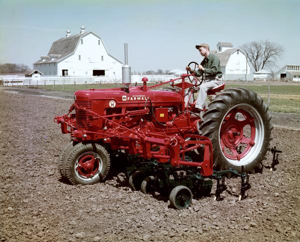 View towards a man cultivating a field with a Farmall Super H tractor and cultivator with farm buildings in the background. The photograph may have been taken at International Harvester's experimental farm at Hinsdale, Illinois.