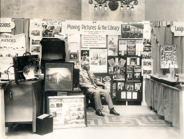 Man sitting in an exhibit of International Harvester films at an American Library Association Conference. The exhibit was titled: "Moving Pictures and the Library" and included photographs, text and film projection equipment.