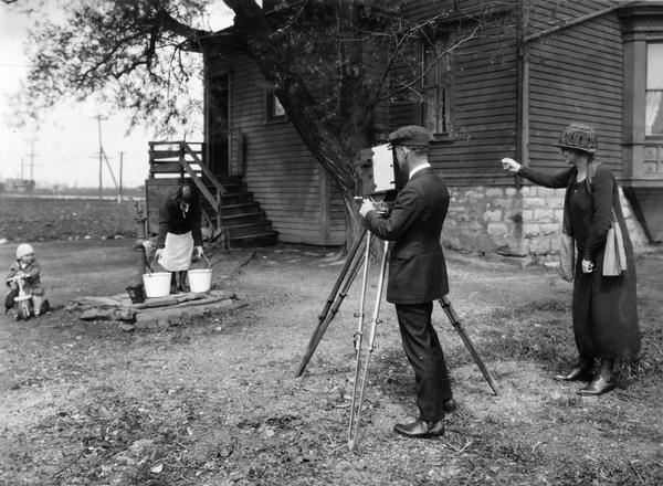 Mr. Hyde and Miss Wigent filming a woman and child while the woman is filling pails with water from a hand-pump. The original caption reads: "Taking footage for Home Economics Reel."