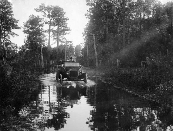 Three men are driving on a flooded rural road. Original caption reads: "In going from Fairhope, Ala. to Point Clear, Ala. the road in many places was found to be serving the double purpose of both road and river." The image was taken for International Harvester's Agricultural Extension Department to document examples of poor roads.
